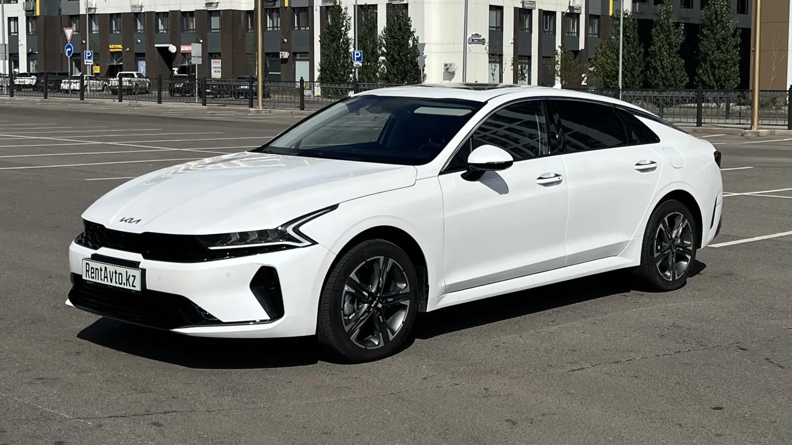 Белая KIA K5 показана спереди на асфальтовой площадке, автомобиль подают с персональным водителем для поездок по городу